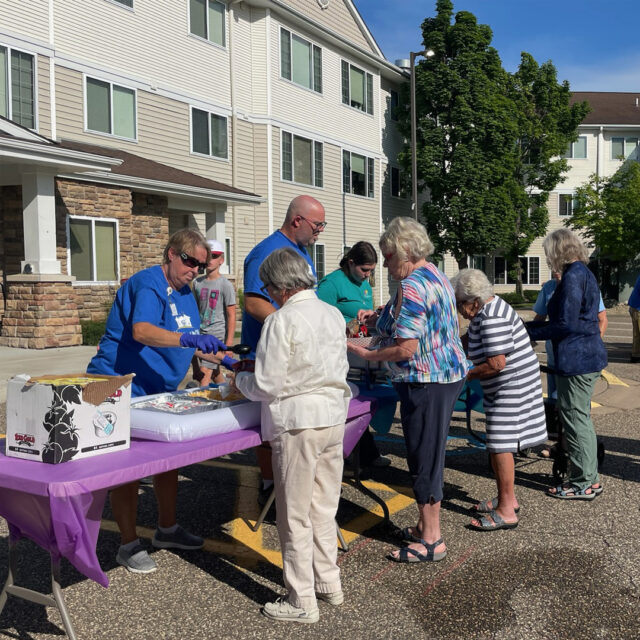 A group of residents and guests lining up at an outdoor food station during the "Night to Unite" event at Cornerstone Assisted Living. Staff members, wearing blue shirts and gloves, are serving walking tacos and other refreshments to the residents. The event is taking place in front of a building on a sunny day, with a festive atmosphere.