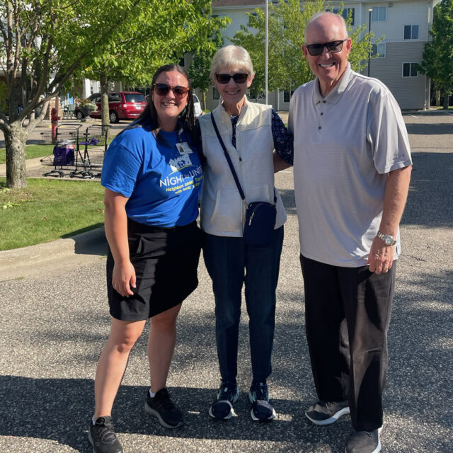 A staff member wearing a blue "Night to Unite" t-shirt stands smiling with two people during the "Night to Unite" event at Cornerstone Assisted Living. The trio is posing together in a parking lot, with trees and the building visible in the background on a sunny day.