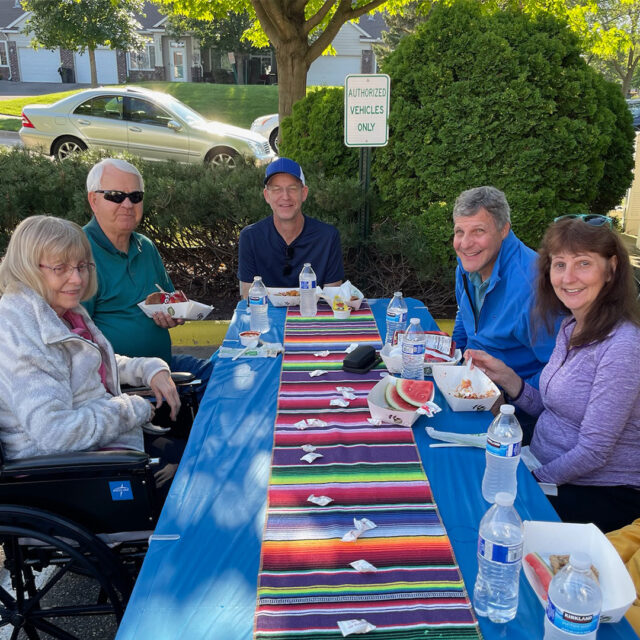 Residents and guests, including a man, a woman, and an elderly lady, sitting at a table enjoying walking tacos and other refreshments. The atmosphere is lively, with attendees in the background and trees providing shade.