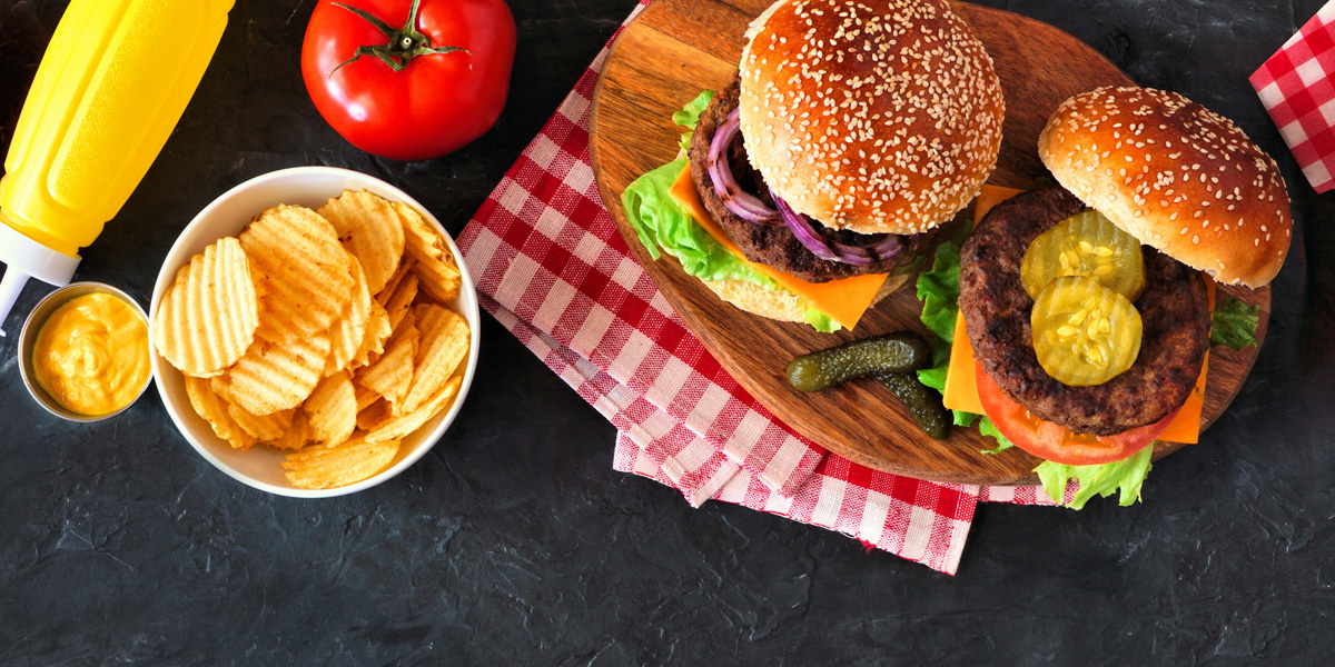 Overhead view of two cheeseburgers on a wooden board with potato chips, mustard, and picnic-style napkins on a dark tabletop.