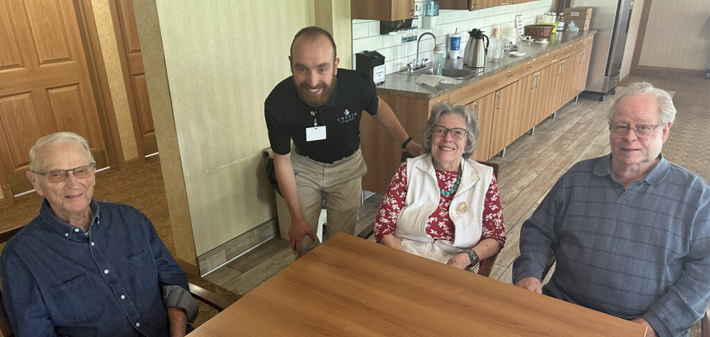 Chaplain at Cornerstone Assisted Living visiting with three residents seated around a table in a community common area with kitchen cabinetry and countertops in the background.